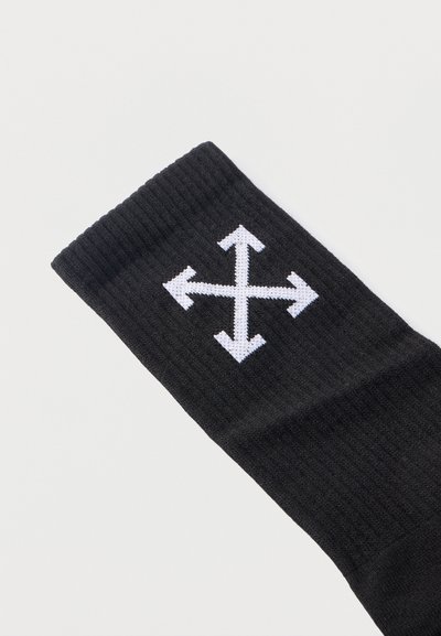 Chaussette noire côtelée avec un symbole de flèche blanche à quatre directions cousu près du haut du bord sur un fond clair uni.