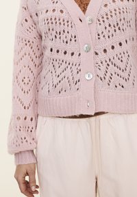 Light pink knitted cardigan with openwork pattern, featuring button closure and ribbed hem. Paired with light-colored fabric pants.