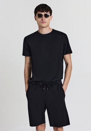Schwarzes T-Shirt mit kurzen Ärmeln, kombiniert mit schwarzen Shorts mit elastischem Bund und Kordelzug. Klare runde Sonnenbrillen runden das Outfit ab.