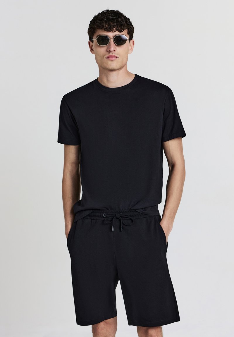 Schwarzes T-Shirt mit kurzen Ärmeln, kombiniert mit schwarzen Shorts mit elastischem Bund und Kordelzug. Klare runde Sonnenbrillen runden das Outfit ab.