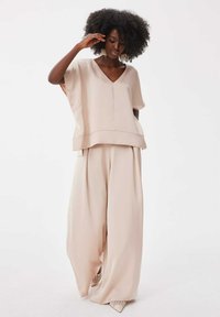 Beige oversized top met een V-hals en brede mouwen, gecombineerd met losse beige broeken. De outfit heeft een gladde textuur en subtiele naden.