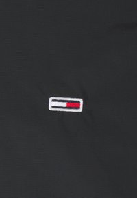Malé obdélníkové logo s bílými, černými a červenými vodorovnými pruhy vyšívané na černém textilním pozadí.