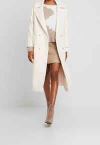 Long manteau crème en mélange de laine avec larges revers, boutonné croisé, associé à un pull texturé beige et blanc avec une jupe, et des bottines à motifs.