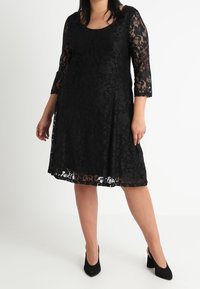 Svart spetsklänning med trekvartsärmar, rundad halsringning och flödande A-linjeform; genomskinliga blommönster med en fodrad underdel.