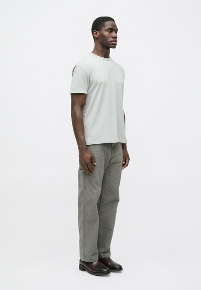 Svetlosivé bavlnené tričko v kombinácii s olivovo zelenými cargo nohavicami a tmavohnedými čižmami. Model stojí v profile na jednoduchom pozadí.