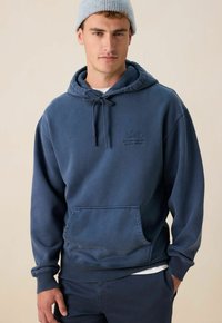 Mann in einem blauen Hoodie mit Fronttasche und Kordelz ügen, heller blauer Strickmütze und dunklen Hosen, der vor einem schlichten Hintergrund steht.