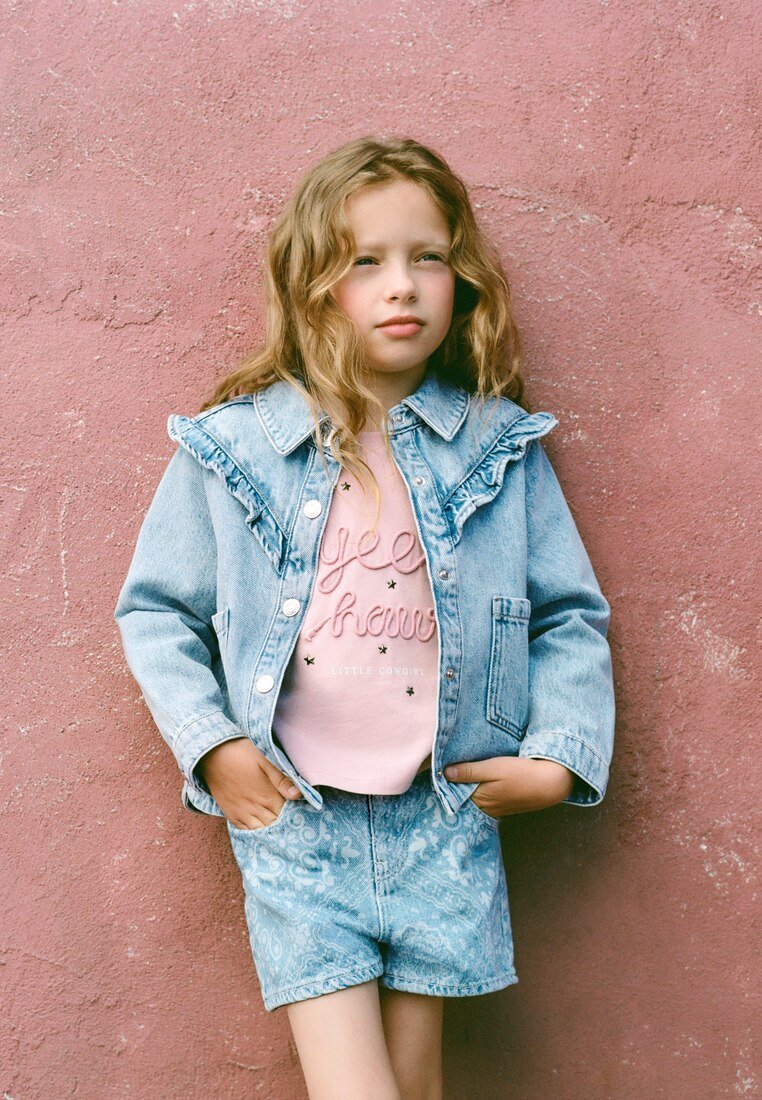 Jeune fille aux cheveux blonds ondulés portant une veste en jean bleue et un short, ainsi qu'un t-shirt rose, se tenant devant un mur rose texturé.