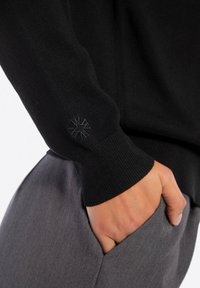 Manica di maglione nero a maglia con polsino a coste e dettaglio del logo ricamato. Pantaloni grigi con una mano inserita nella tasca laterale.
