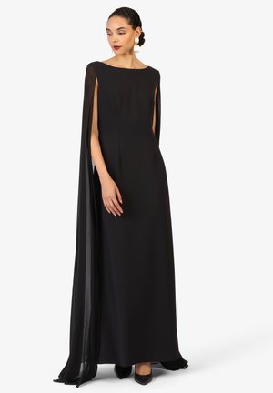 Robe noire longue avec un corsage ajusté, décolleté bateau et manches en cape transparentes. La texture lisse complète des escarpins à bout pointu.