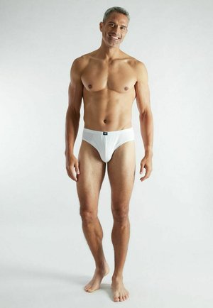Smiling fit man standing barefoot wearing white briefs against a plain light background.