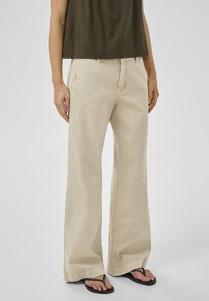 Személy bézs színű, bő szárú nadrágot, fekete papucsot és sötét olívazöld, rövid felsőt visel, egyszínű háttér előtt áll.