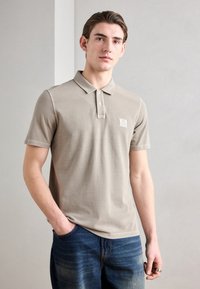 Beige polo-skjorte med to-knapps knapplukking og korte ermer, kombinert med blå denimjeans. Har en liten logo på venstre bryst.