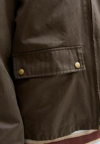 Giacca marrone con una texture lucida, dotata di una tasca frontale piatta fissata da due bottoni dorati. Chiusura con zip su un lato.