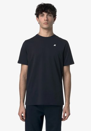 Mladý muž s kučeravými vlasmi, ktorý nosí jednoduché čierne tričko s krátkym rukávom a malým bielym logom na ľavej strane hrudníka, stojaci pred bielym pozadím.