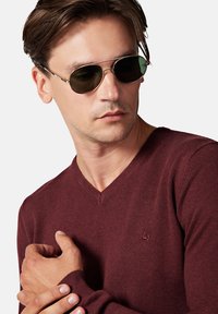 Burgundowy sweter z dekoltem w serek, z prążkowanymi mankietami i małym tonowym logo. Model nosi zielone okulary przeciwsłoneczne, prezentując casualowy wygląd.