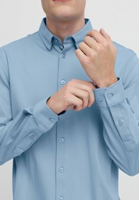 Camisa de manga larga azul claro con cuello abotonado y puños abotonados. Tejido suave con un corte entallado. Cuatro botones en la parte delantera.