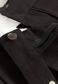 Gros plan d’un pantalon à carreaux foncé montrant un bouton, une fermeture éclair et les détails de la ceinture sur un tissu texturé.