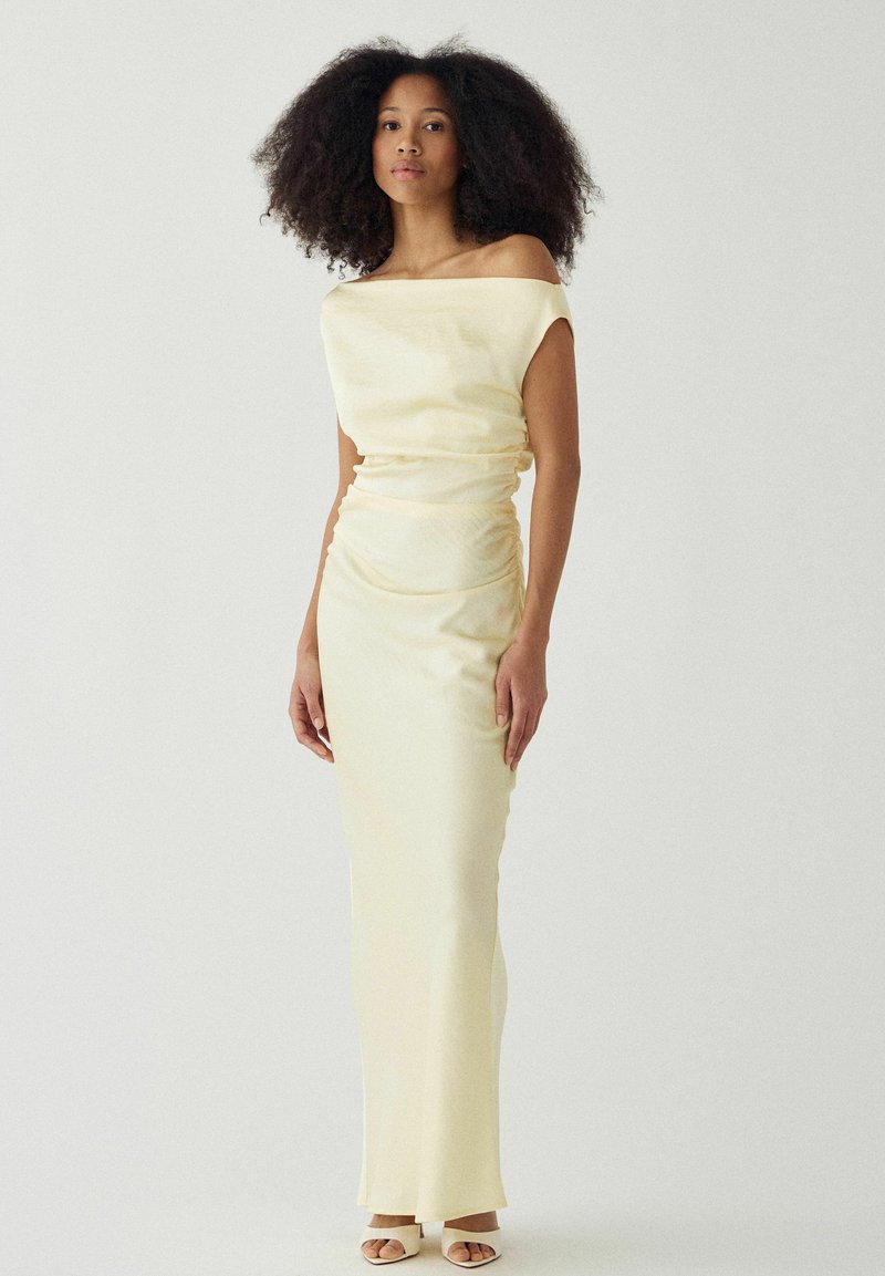 Femme aux cheveux bouclés et volumineux portant une robe froncée jaune pâle à épaules dénudées et des talons blancs ouverts, posant devant un fond uni.
