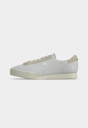 Λευκό sneaker χαμηλού ύψους με μπεζ σόλα και λεπτομέρειες, με μεταλλικό λογότυπο στο πλάι και κλείσιμο με κορδόνια σε απλό ανοιχτό φόντο.