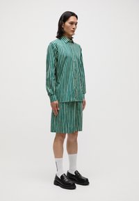 Cămașă cu dungi verzi și bej, cu butoane, asortată cu o fustă corespunzătoare. Modelul poartă pantofi negri și șosete albe, evidențiind un aspect casual.