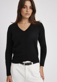 Pull noir à manches longues avec un décolleté en V, fabriqué dans un matériau doux. Porté avec une ceinture blanche et un pantalon clair.