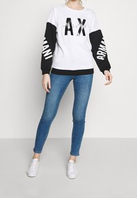 Weißer Pullover mit schwarzen Ärmeln, mit "AX" und "ARMANI" Grafiken. Kombiniert mit blauen Skinny Jeans und weißen Sneakers.