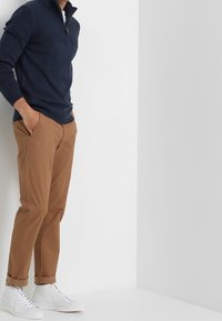 Pull-over en maille bleu marine avec col à fermeture éclair partielle, associé à un pantalon chino beige et des baskets blanches. Texture lisse et coupe ajustée avec revers aux manches.