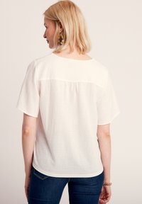 Top bianco a maniche corte con tessuto texturizzato, scollatura rotonda e dettaglio plissettato sul retro. Abbinato a jeans blu scuri e a un bracciale.