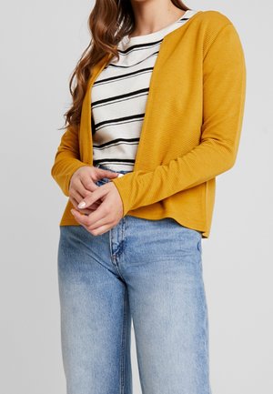 Femme portant un cardigan ouvert jaune moutarde sur une chemise rayée blanche et noire avec un jean taille haute bleu clair sur un fond gris.