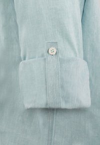 Puño de camisa de lino azul claro con un solo botón, que presenta un diseño enrollado y una textura sutil. Detalle de costura visible a lo largo de las costuras.
