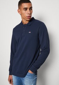 Polo a maniche lunghe blu navy con colletto, due bottoni e un piccolo logo sul petto; abbinato a jeans in denim chiaro con effetti usurati.