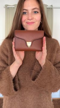 Brūna teksturēta ādas maks ar zelta detaļām, kas veidots ar atloku un izliektu dizainu. Tur rokās uz neitrāla fona.