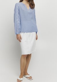 Vaaleansininen puuvillapusero V-pääntiellä ja kirjailuilla hihoissa, yhdistettynä valkoiseen farkkuhameeseen ja valkoisiin liukusandaaleihin.