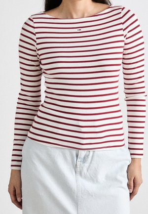 Une femme portant une chemise blanche ajustée à manches longues avec des rayures rouges horizontalement et une jupe en denim blanche avec des poches.