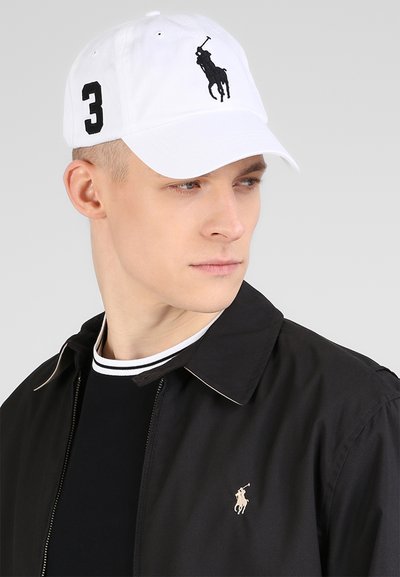 Balts beisbola cepure ar melnu polo logotipu un izšūtu numuru "3" sānos, izgatavota no kokvilnas materiāla, ar izliekto malu, valkāta kopā ar melnu jaku.