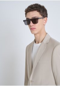 Blazer beige ajusté avec une fermeture à un bouton et une poche poitrine, porté sur un t-shirt blanc. Des lunettes de soleil noires complètent le look.