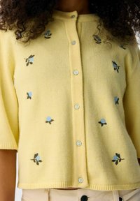 Geel kortemouw cardigan met geborduurde blauwe bloemmotieven. Heeft een knoopsluiting aan de voorkant met lichtblauwe knopen en een zachte textuur.