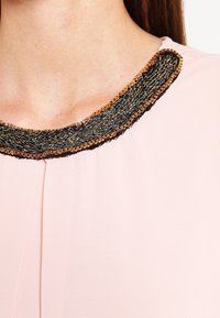 Blusa rosa claro con escote redondo, que presenta un ribete texturizado en negro y dorado en el collar y un acabado de tela suave.