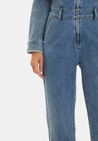 Diminutivo tuta in denim di un blu chiaro con vita aderente, leggero lucido, e dettagli ai polsini che presentano una piccola rifinitura di borchie lungo i bordi delle maniche.