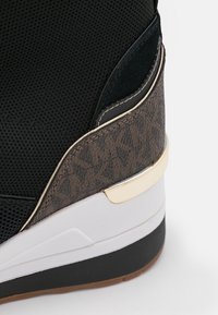 Zapatilla negra con parte superior de malla, panel marrón con patrón texturizado, acento dorado en el talón, mediasuela blanca y suela exterior de caucho negra.