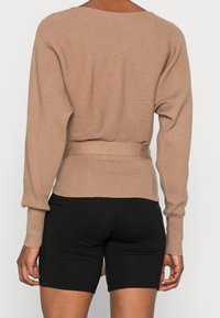 Light brown ribbed sweater with a loose fit and belted waist, long sleeves, and a scooped back neckline, paired with black shorts.