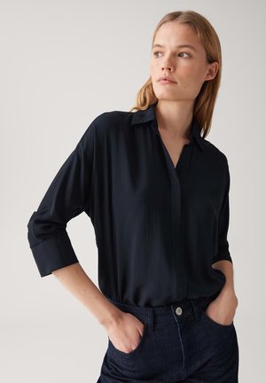 Marineblaue Bluse aus leichtem, glattem Material; mit Kragen, durchgehender Knopfleiste und Dreiviertelärmeln, kombiniert mit dunklen Jeans.