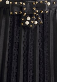 Falda negra plisada hecha de un material de cuero texturizado, adornada con tachuelas doradas y acentos de perlas. Detalle de lazo en la cintura.