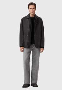Jeune homme portant une veste en daim noire sur un pull noir, un jean gris clair et des chaussures noires, se tenant contre un fond clair.