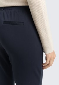 Navy stretchbyxor med en stilren design, med en bakficka och mjuk textur. En ljusrandig långärmad topp är delvis synlig.