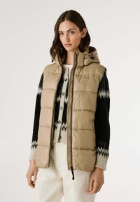 Beige gewatteerde vest met capuchon, voorzien van een rits aan de voorkant en zwarte gebreide mouwen met een geometrisch patroon. Draag het over een crèmekleurige trui.