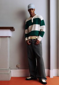 Grün-weiß gestreifter Pullover mit Kragen, graue weite Jeans und schwarz-weiße Schuhe. Hellblaues Cap mit Logo, silberner Schmuck.