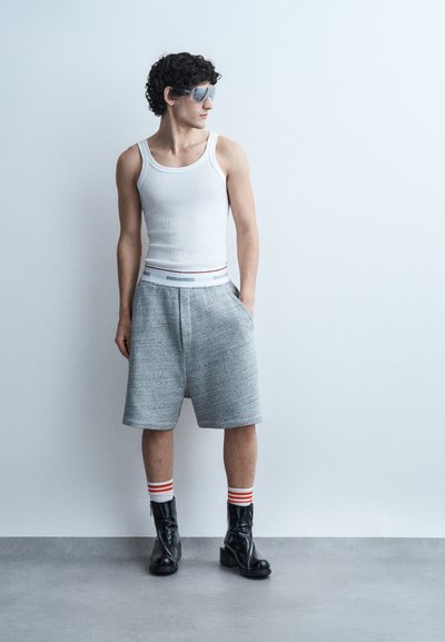 Νέος άνδρας με λευκό αμάνικο μπλουζάκι, γκρι σορτς, μαύρα μποτάκια, ριγέ καλτσάκια και ανακλαστικά γυαλιά ηλίου στέκεται δίπλα σε λευκό τοίχο.