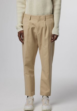Pantalons beige ajustés avec plis, présentant une texture lisse, des passants de ceinture et une coupe décontractée, associés à des baskets blanches.