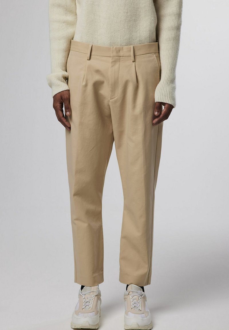 Pantalons beige ajustés avec plis, présentant une texture lisse, des passants de ceinture et une coupe décontractée, associés à des baskets blanches.